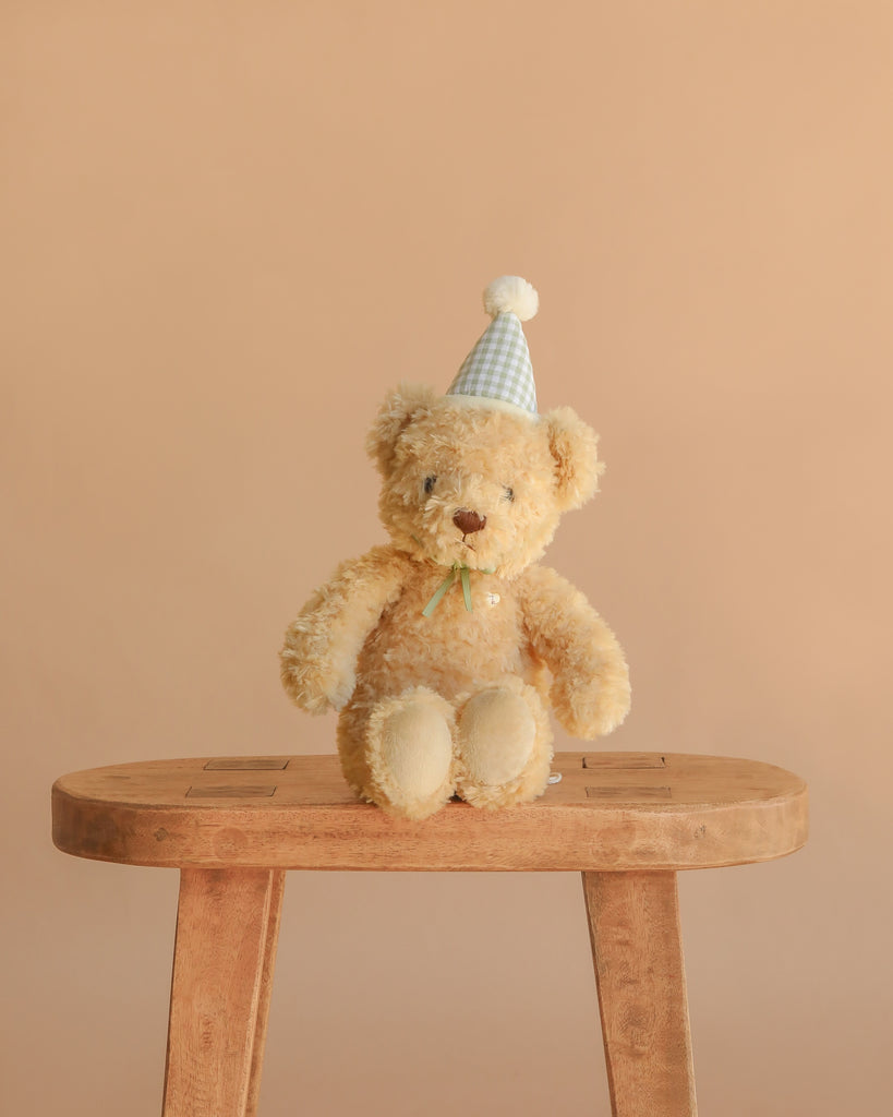 The Snuggle Bunnies - Birthday Arthur - Gingham is a plush toy bear in a blue and white party hat, seated on a wooden stool against a beige background—an ideal keepsake gift.
