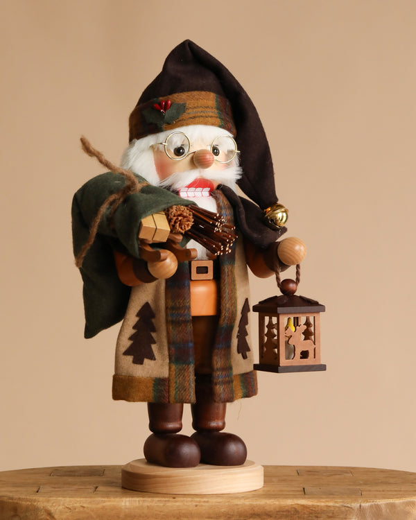 The Christian Ulbricht Collectible Wooden Nutcracker - Natural Woodsy Santa With Lantern stands on a round wooden base, wearing plaid clothes and boots, holding a green sack, firewood, and lantern. Made in Germany.