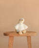 Snuggle Bunnies - Rose The Duck, a white plush toy with yellow beak and feet, sits on a wooden stool against a beige background—an ideal super soft companion for cozy moments.