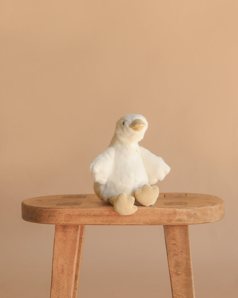 Snuggle Bunnies - Rose The Duck, a white plush toy with yellow beak and feet, sits on a wooden stool against a beige background—an ideal super soft companion for cozy moments.