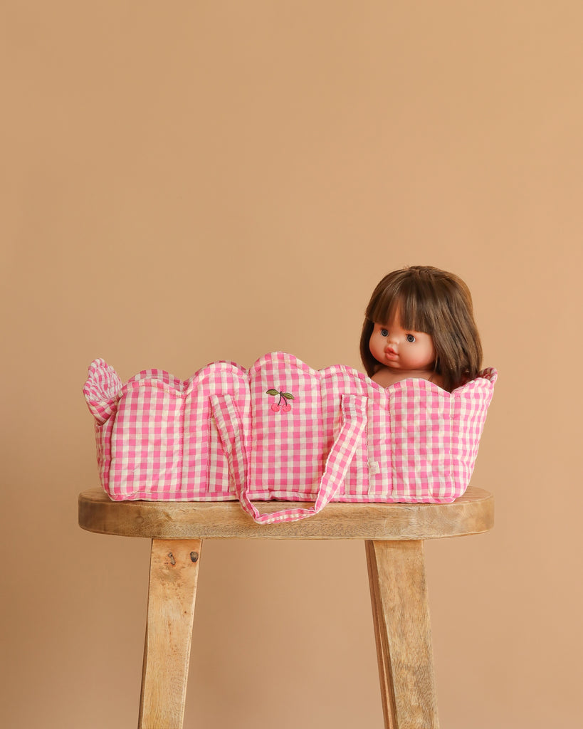 A doll with brown hair sits partially inside the Konges Sloejd Doll Lift - Bubblegum Check, a pink and white gingham-patterned OEKO-TEX cotton carrier, resting on a wooden stool against a plain beige background.
