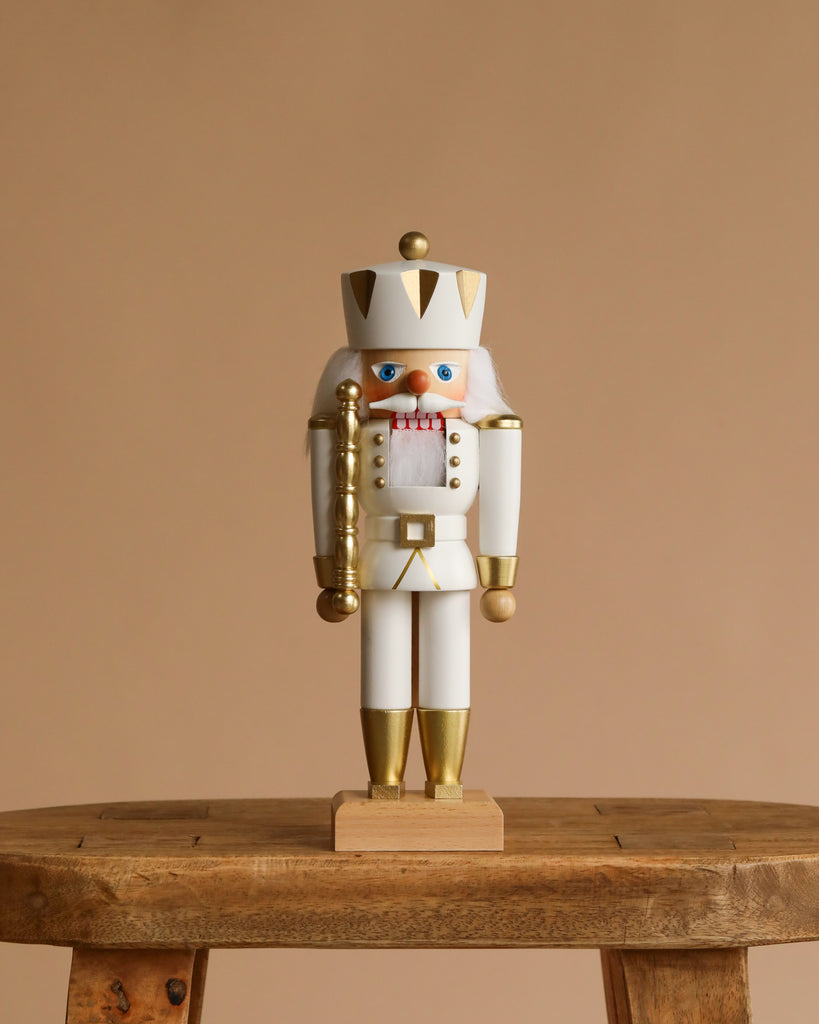 The Christian Ulbricht Collectible Wooden Nutcracker - King (White and Gold) stands upright on a rustic wooden stool, crowned and mustached, against a plain beige background.