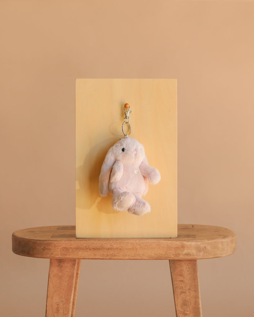 The Snuggle Bunnies - Penelope Keyring - Blossom, a soft light pink plush bunny, hangs on a hook attached to a wooden board set upright on a simple stool against a plain beige background.