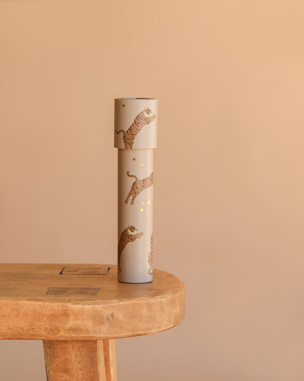 The Konges Sloejd Kaleidoscope - Tiger, a beige cylinder adorned with tiger illustrations, gold stars, and colorful pearls, stands upright on a light wooden table against a plain beige background.
