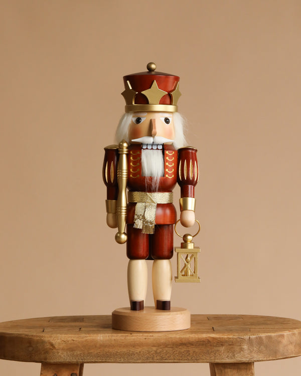 The Christian Ulbricht Collectible Wooden Nutcracker - Prince (Natural and Gold), made in Germany, stands on a wooden table against a plain beige background, wearing a crown and holding a staff and lantern.