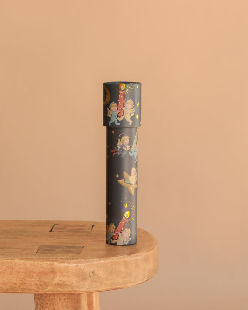 The Konges Sloejd Kaleidoscope - Lucia, a black cylinder adorned with playful children and star illustrations, stands upright on a light wooden table against a solid beige background.
