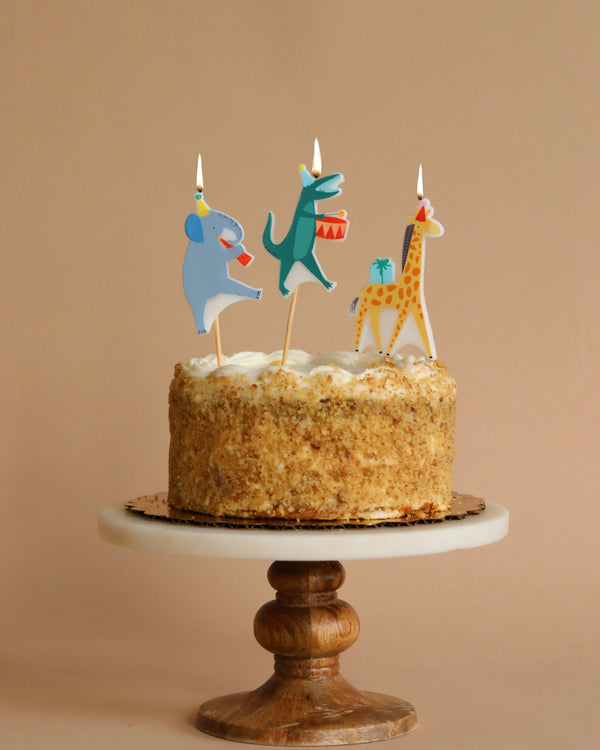 A birthday party cake on a wooden stand topped with cream frosting and colorful animal toppers including an elephant, fox, and giraffe, each holding a gift, with Meri Meri Animal Parade Candles.