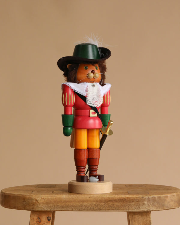 The Christian Ulbricht Collectible Wooden Nutcracker - Puss 'n Boots wears a black hat, green gloves, red coat, yellow pants, and white lace collar, standing on a small wooden stool against a beige background.