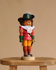 The Christian Ulbricht Collectible Wooden Nutcracker - Puss 'n Boots wears a black hat, green gloves, red coat, yellow pants, and white lace collar, standing on a small wooden stool against a beige background.