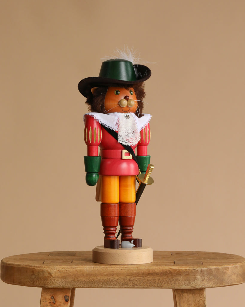 The Christian Ulbricht Collectible Wooden Nutcracker - Puss 'n Boots wears a black hat, green gloves, red coat, yellow pants, and white lace collar, standing on a small wooden stool against a beige background.