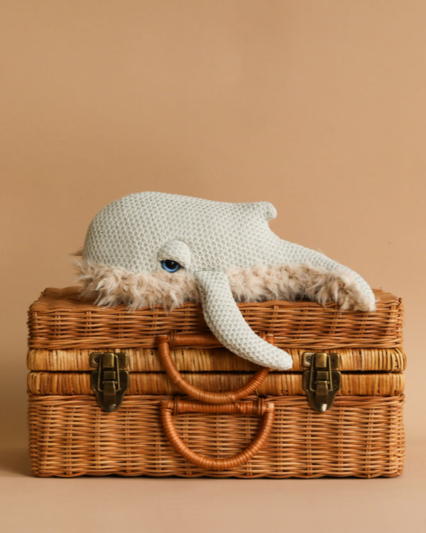 A BigStuffed Mini Grandma Whale, crafted from quality materials with a light blue, textured body and a single visible eye, rests on top of a wicker suitcase with brass locks and a leather handle. This charming scene, featuring a mini whale comforter beside it, is set against a beige background.