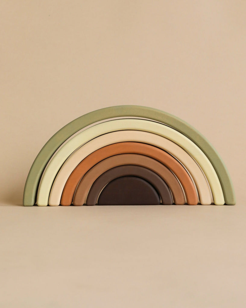 A minimalist, multicolored stackable wooden rainbow toy with seven arches arranged by size. Handmade and painted with non-toxic paint in earthy tones like green, beige, brown, and cream, the Handmade Rainbow Stacker - Olive sits beautifully against a neutral beige background.
