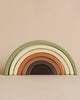 A Handmade Rainbow Stacker - Olive featuring seven arch segments in muted earth tones, ranging from light green, cream, tan, and brown. Made with non-toxic paint, the piece is set against a plain beige background.