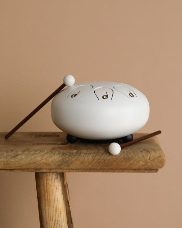 The Steel Tongue Drum, a white musical instrument with black feet, rests on a rustic wooden stool next to two brown mallets with white tips, all against a beige background.