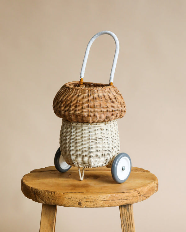A whimsical Olli Ella Rattan Mushroom Luggy - Natural shaped like a pot and mounted on small wheels, placed on a rustic wooden stool against a neutral beige backdrop.