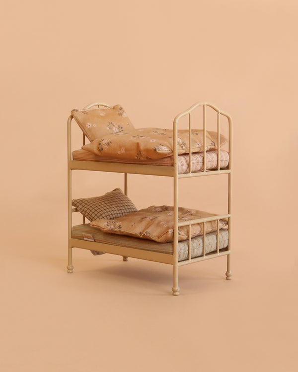 A small, cream-colored Maileg Bunk Bed with floral bedding and pillows is set against a solid beige background. Ideal as a sleepover toy, this miniature bed from Mouse Hole Farmhouse is perfectly sized for dolls.