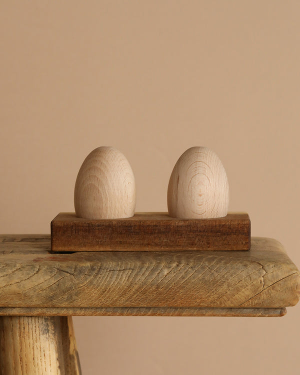 The Wooden Egg Shakers with Stand features two smooth, unfinished shakers on a small wooden stand—ideal for encouraging developmental milestones through natural music play, beautifully set against a plain beige background.