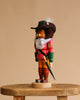 The Christian Ulbricht Collectible Wooden Nutcracker - Puss 'n Boots, dressed as a musketeer with a feathered hat, lace collar, red and green outfit, and sword, stands on a wooden stool against a plain beige background.