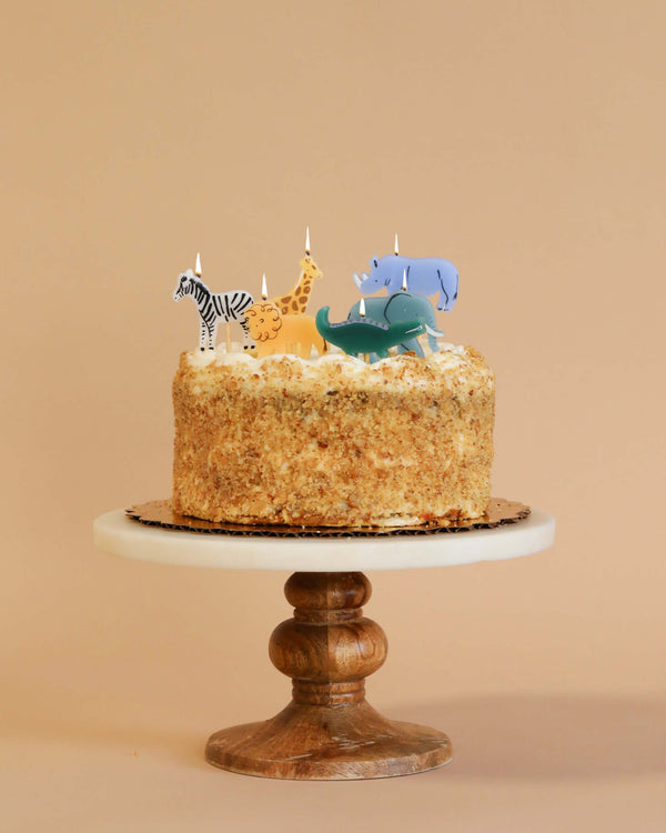 A birthday cake with a crumb coating on a wooden stand, decorated with Meri Meri Jungle Animal Candles including a zebra, giraffe, and elephant. The background is a soft beige tone.