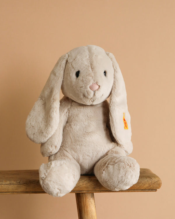 The Steiff Hoppie Bunny Stuffed Animal, a soft, light beige plush with long floppy ears and black eyes, sits on a wooden stool against a plain beige background.