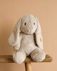 The Steiff Hoppie Bunny Stuffed Animal, a soft, light beige plush with long floppy ears and black eyes, sits on a wooden stool against a plain beige background.