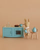 The Maileg Blue Kitchen with Rabbit Set features a stuffed rabbit in a dress holding a blue cup, sitting on a wooden stool beside the blue kitchen set and teapot, all against a beige background.