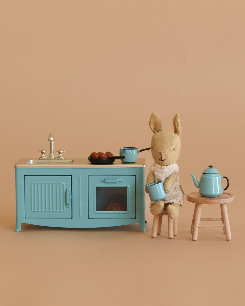 The Maileg Blue Kitchen with Rabbit Set features a stuffed rabbit in a dress holding a blue cup, sitting on a wooden stool beside the blue kitchen set and teapot, all against a beige background.
