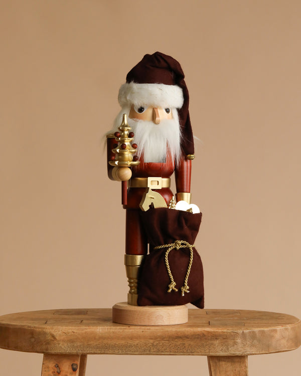 The Christian Ulbricht Collectible Wooden Nutcracker - Santa (Natural and Gold) features Santa in a brown hat and coat, holding a gold staff and brown sack, standing on a wooden stool against a beige background.