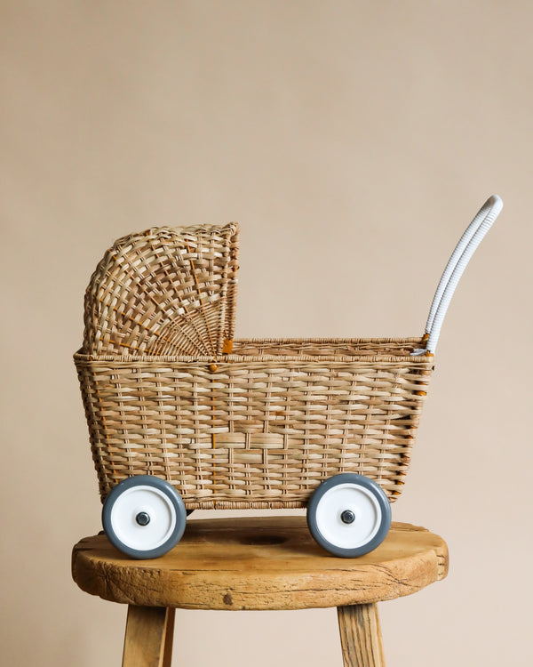 A hand-woven Olli Ella Rattan Doll Stroller with two large blue wheels sits on a wooden stool against a neutral beige background. Its handle is wrapped with white material for a comfortable grip.