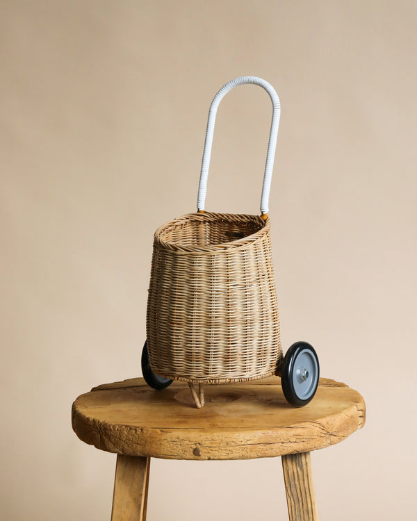 A hand-woven Olli Ella Rattan Luggy on wheels with a white curved handle, standing on an old wooden stool against a plain beige background.