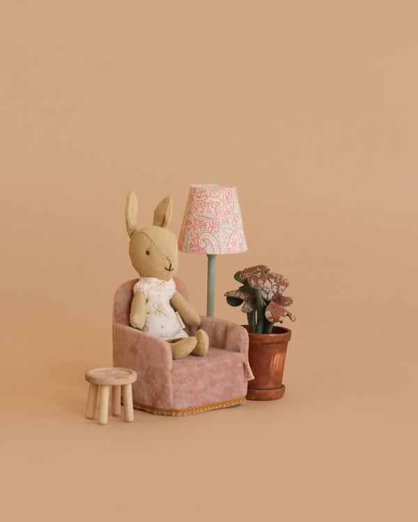 The Maileg Little Rabbit’s Reading Nook features a stuffed bunny on a plush pink armchair beside a wooden stool, Vintage Floor Lamp with patterned shade, and potted plant, set against a simple beige background.
