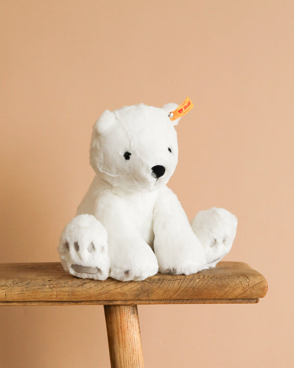 The Steiff Lasse Polar Bear Stuffed Animal - 11", crafted from ultra-soft white fabric and featuring a yellow ear tag, sits on a wooden bench against a plain beige background.