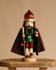 The Christian Ulbricht Collectible Wooden Nutcracker - Forest King, made in Germany, stands on a wooden stool in a brown cloak, green shirt, red pants, and gold crown, holding a staff with mushrooms at its feet against a beige background.