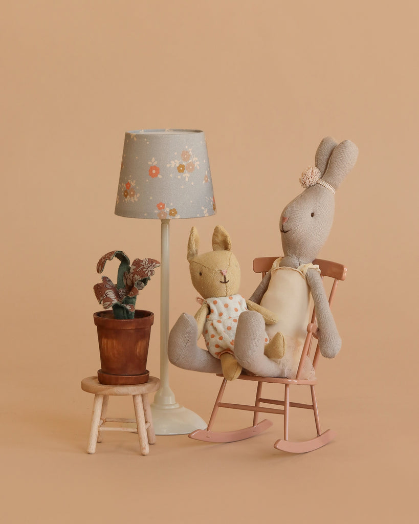 The Maileg Little Rabbit’s Rocking Chair Set features two plush rabbits on a pink rocking chair, a nearby small stool with a potted plant, and a white lamp with a flower-patterned shade, all set against a beige background.