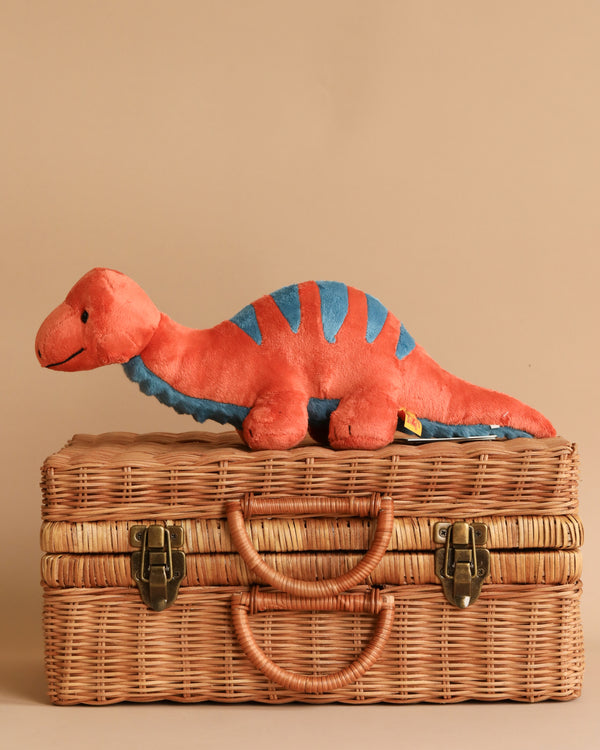 A Steiff, Bronko Brontosaurus Dinosaur Plush Stuffed Toy, 11 Inches shaped in vibrant red and blue lies atop a closed wicker basket with leather handles and brass latches. The background is a plain beige color, creating a simple and warm setting.
