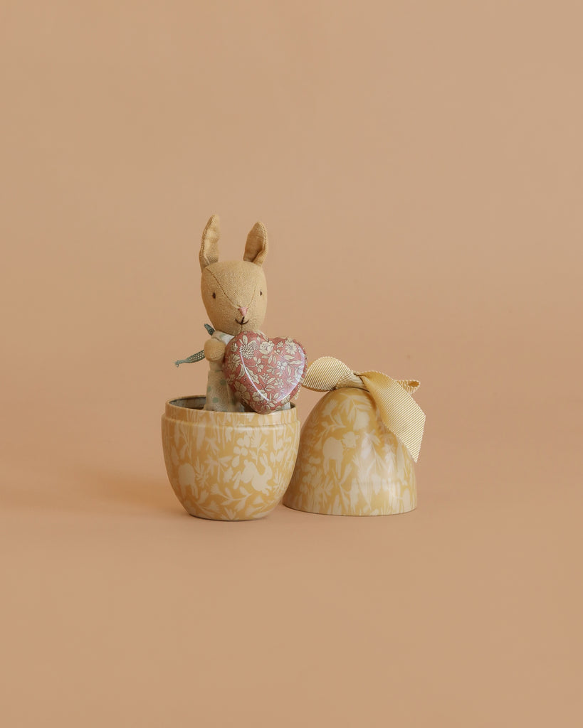 The Maileg Easter Egg with Micro Rabbit & Heart Set features a small stuffed bunny holding a pink heart inside an egg-shaped container with lid and yellow ribbon, displayed against a plain beige background.