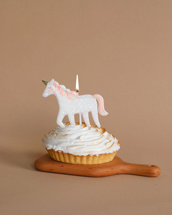 A Meri Meri Unicorn Glitter Candle lit on top of a creamy frosted cupcake, presented on a wooden paddle. The background is a smooth, plain tan color.
