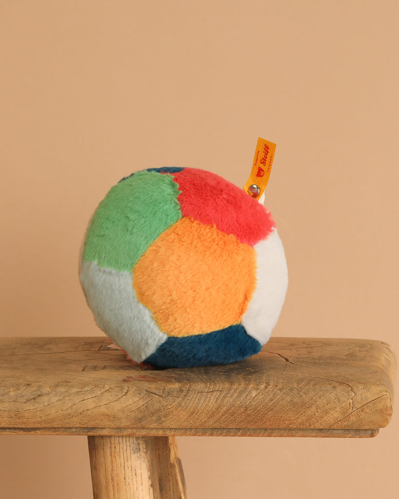 The Steiff Multicolored Plush Ball with Rattle, made from soft, fuzzy fabric for easy gripping, features green, red, orange, blue, and white segments and rests on a rustic wooden stool.