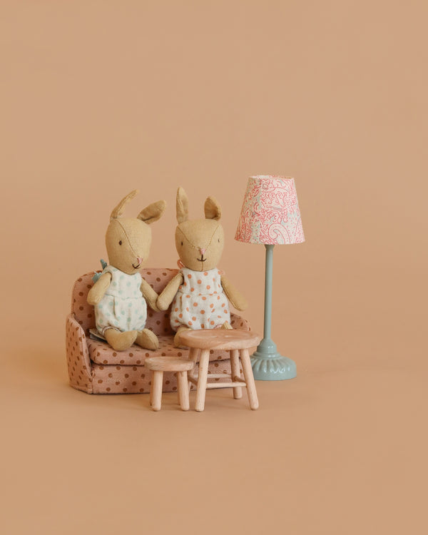 Two stuffed bunny dolls sit on the Maileg Little Rabbit’s Living Room Set, which includes a polka-dot sofa bed, blue floor lamp, wooden table, and stool, all arranged against a plain beige background.