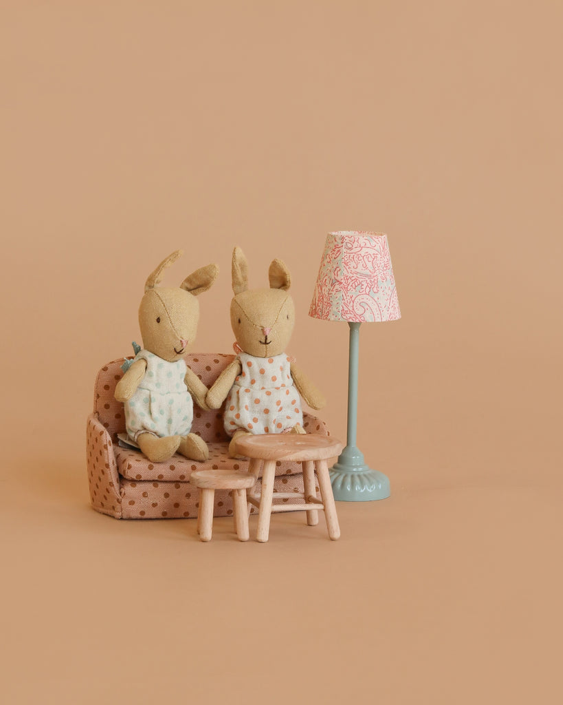 Two stuffed bunny dolls sit on the Maileg Little Rabbit’s Living Room Set, which includes a polka-dot sofa bed, blue floor lamp, wooden table, and stool, all arranged against a plain beige background.