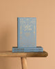 A Luxe Edition hardcover of "The Great Gatsby" by Fitzgerald, featuring ornate gold detailing, stands upright on a wooden bench with two identical copies beneath it, all set against a plain beige background.