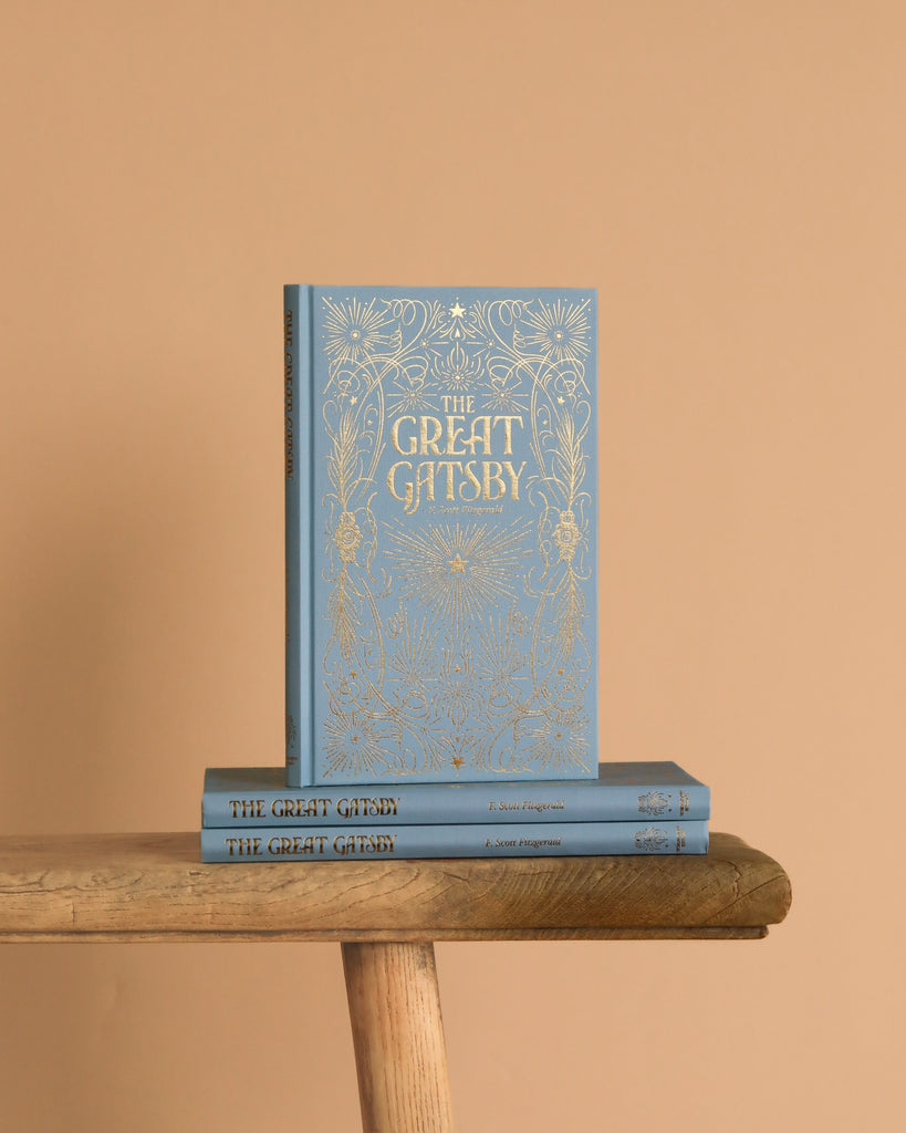 A Luxe Edition hardcover of "The Great Gatsby" by Fitzgerald, featuring ornate gold detailing, stands upright on a wooden bench with two identical copies beneath it, all set against a plain beige background.