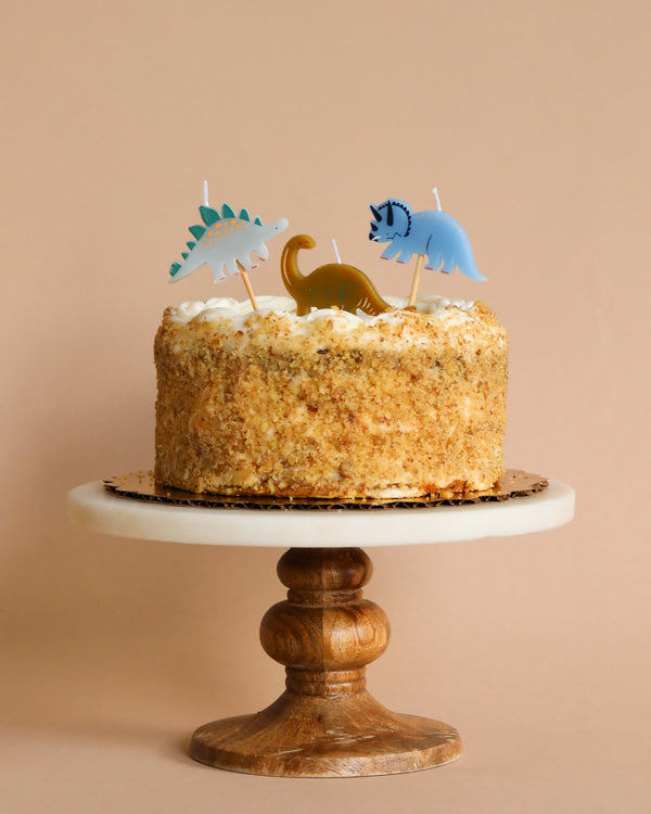A birthday cake adorned with Meri Meri Dinosaur Candles, presented on a wooden stand against a soft peach background.