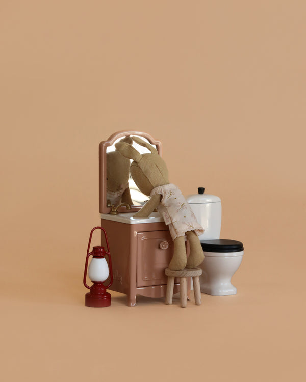 A Maileg Little Rabbit’s Bathroom Set features a mouse in a dress standing on a stool by a sink dresser with mirror, accompanied by a white toilet and red lantern, all arranged on a plain beige background.
