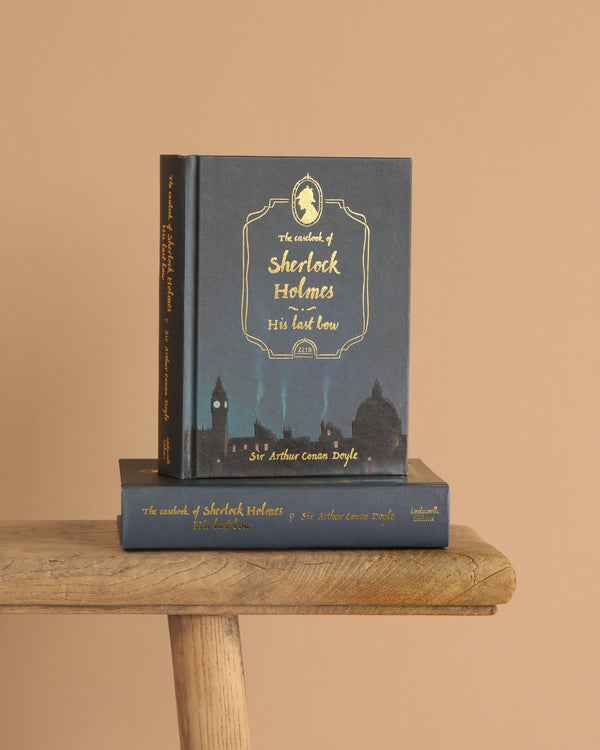 Two copies of "The Casebook of Sherlock Holmes | Collector’s Ed | Hardcover" sit on a wooden bench against a plain beige background, their covers displaying a dark cityscape and bold gold lettering.