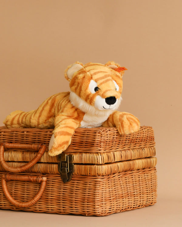 A Steiff, Toni Tiger Stuffed Animal Plush, 12 Inches from the Soft Cuddly Friends collection is laid across a woven, rectangular basket with a clasp in the front. The background is a plain beige color, complementing the tan and orange shades of both the basket and the tiger.