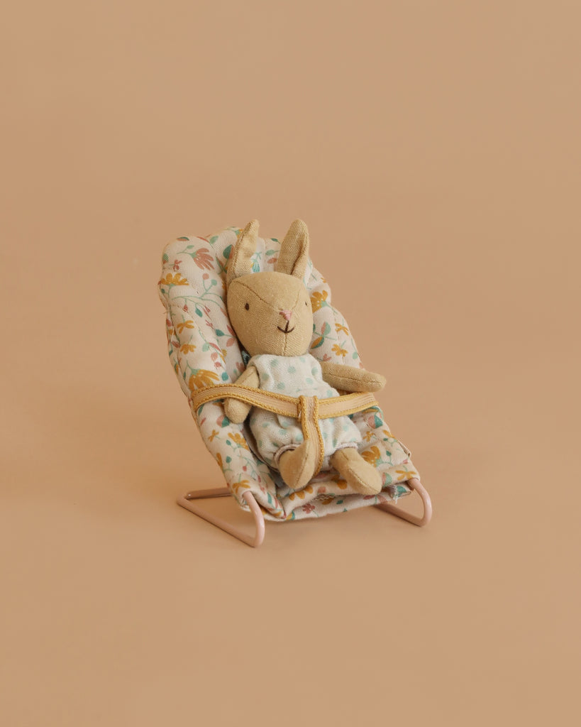 A Maileg Little Rabbit’s Nursery Set features a rabbit in a green dress strapped into a floral baby bouncer, mirroring the cozy comfort of the nursery set, all against a plain beige background.