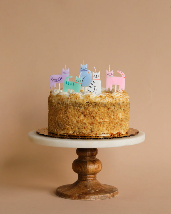 A birthday celebration cake with beige frosting and a crumbly texture, topped with colorful Meri Meri Cat Candles, displayed on a wooden cake stand against a neutral backdrop.