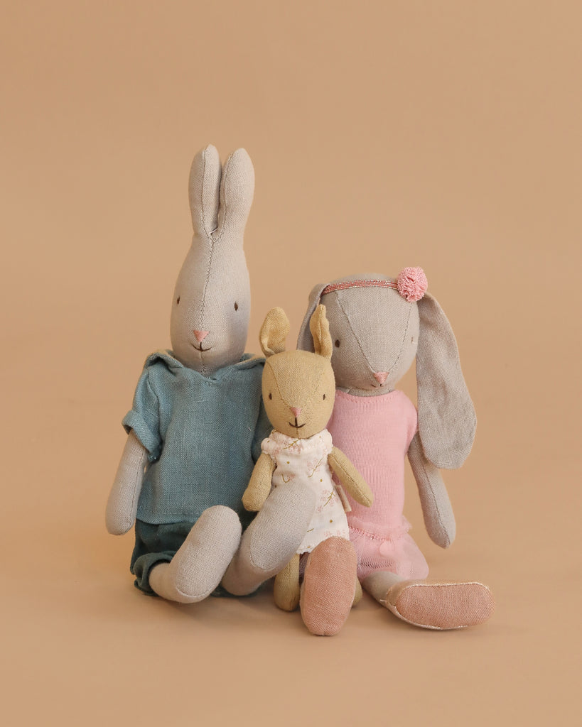 The Maileg Bunny Rabbit Family Set features three stuffed bunnies against a beige background: the largest in blue, the middle in pink with a headband, and the smallest in a white dress with pink dots.