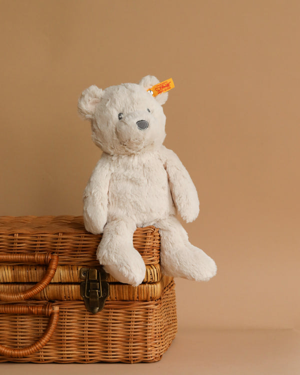 A Steiff, Bearzy Teddy Bear Stuffed Plush For Baby, 11 Inches with a yellow tag on its ear is sitting on top of a closed wicker basket. The background is a solid beige color, creating a warm and cozy atmosphere, perfect for baby's soft skin.